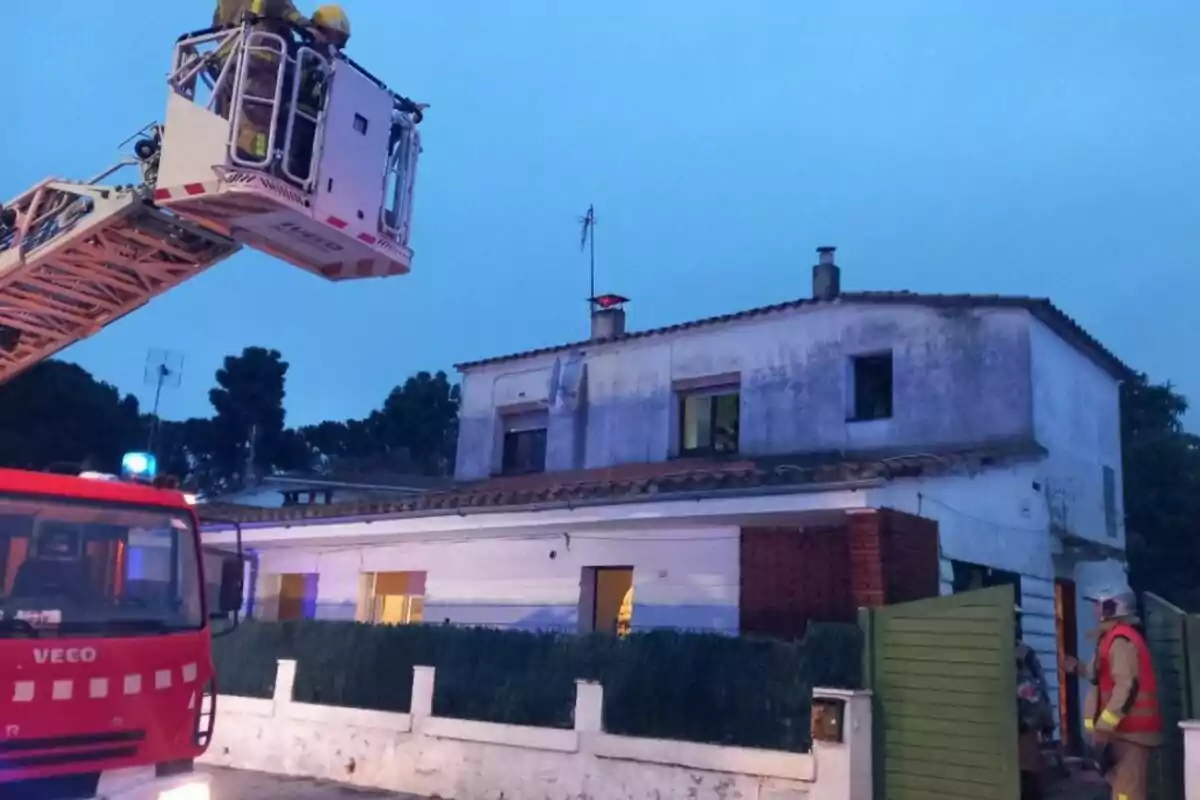 Bombers en una plataforma elevadora davant d'una casa de dos pisos, amb un camió de bombers estacionat al davant i un bomber dret a l'entrada.