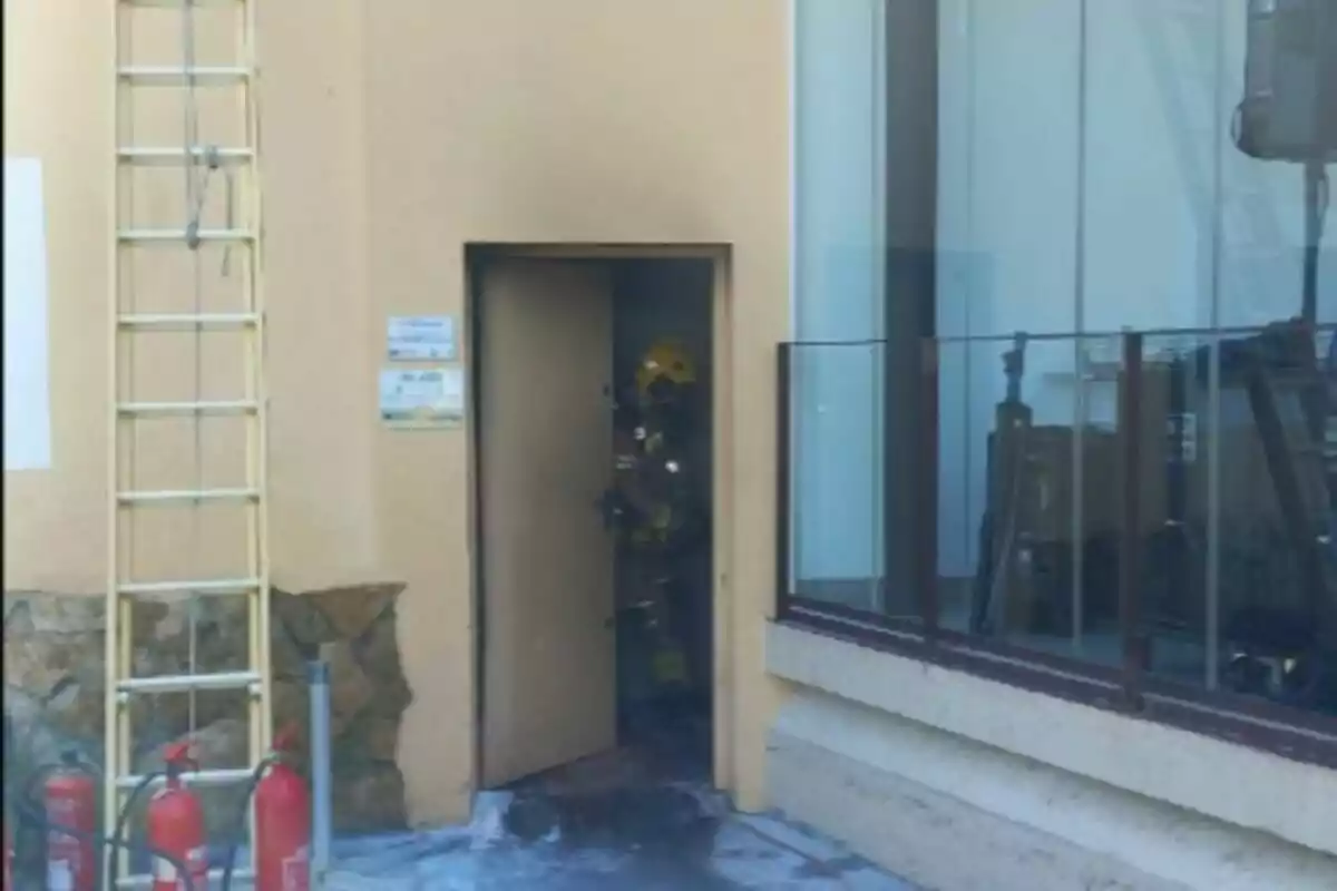 Un bombero con casco amarillo sale de una puerta en un edificio beige, junto a varios extintores y una escalera apoyada en la pared.