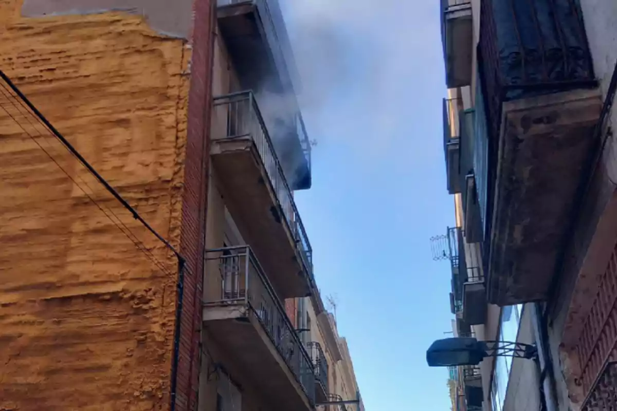 Edificis alts amb balcons i fum sortint d'un d'ells en un carrer estret.
