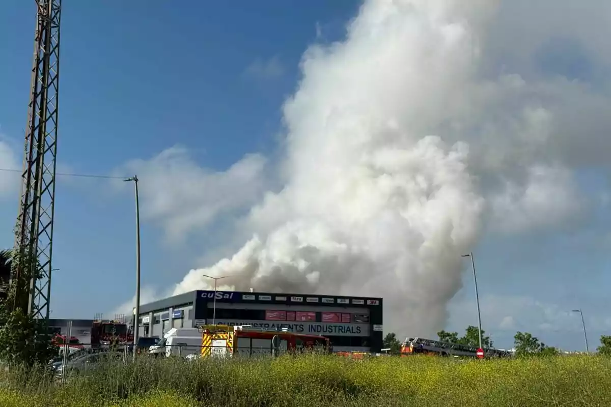 Un edifici industrial amb fum sortint del sostre i vehicles d'emergència al voltant. Un edifici industrial amb fum sortint del sostre i vehicles d'emergència al voltant.