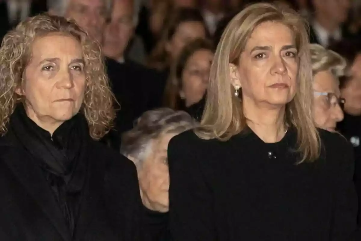 Dos mujeres con expresión seria, vestidas de negro, en un evento con más personas al fondo.