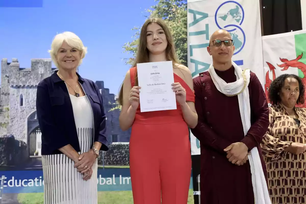 La infanta Sofía sostiene un diploma mientras está de pie entre dos personas adultas, con un fondo de banderas y un castillo. La infanta Sofía sostiene un diploma mientras está de pie entre dos personas adultas, con un fondo de banderas y un castillo.