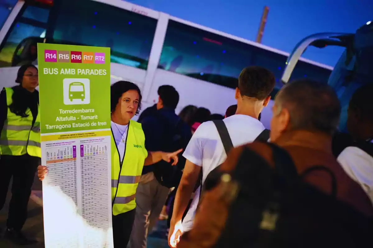 Una treballadora amb armilla reflectora sosté un cartell informatiu de parades d'autobús mentre parla amb un grup de persones davant d'un autobús.