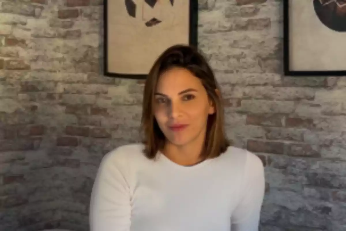 Una persona con cabello castaño y una camiseta blanca está frente a una pared de ladrillos con dos cuadros colgados. Una persona con cabello castaño y una camiseta blanca está frente a una pared de ladrillos con dos cuadros colgados.