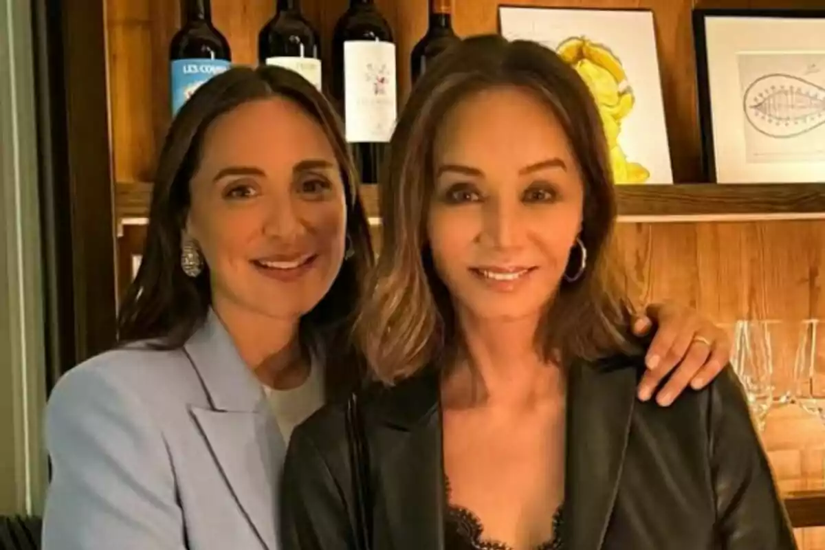 Dos mujeres posando juntas frente a una estantería con botellas de vino y cuadros.