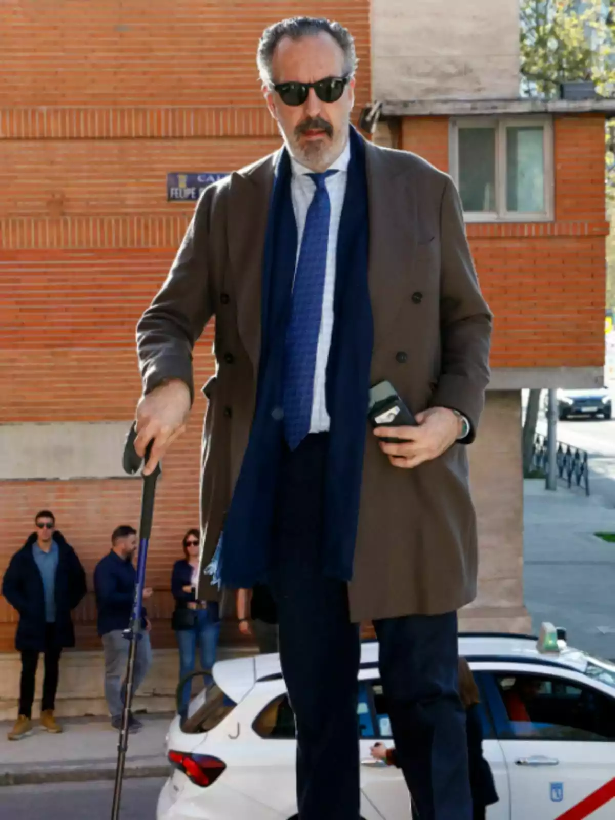 Jaime de Marichalar, wearing sunglasses and carrying a cane, walks down the street in a brown coat and blue tie, with a brick building in the background and several people and a white car behind him.