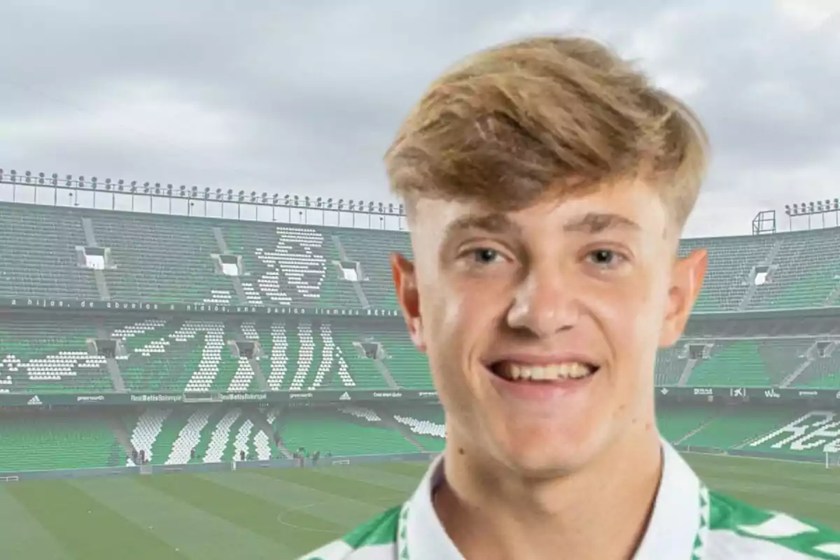 Un joven con camiseta deportiva sonríe frente a un estadio de fútbol con asientos verdes y blancos.