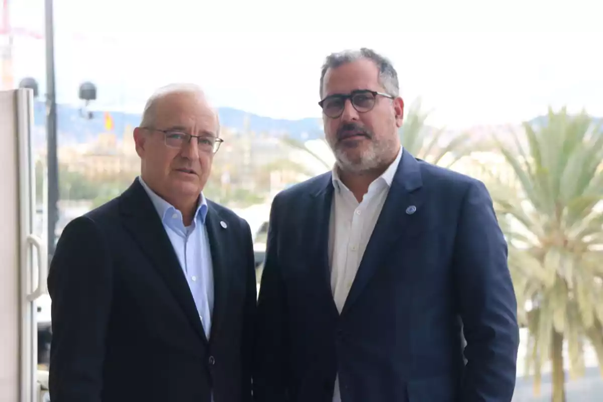 Dos homes de vestit estan junts a l'aire lliure amb palmeres i un paisatge urbà de fons.