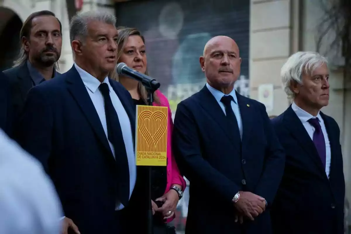 Persones en vestits formals durant un esdeveniment amb un micròfon i un cartell que diu “Diada Nacional de Catalunya 2024”.