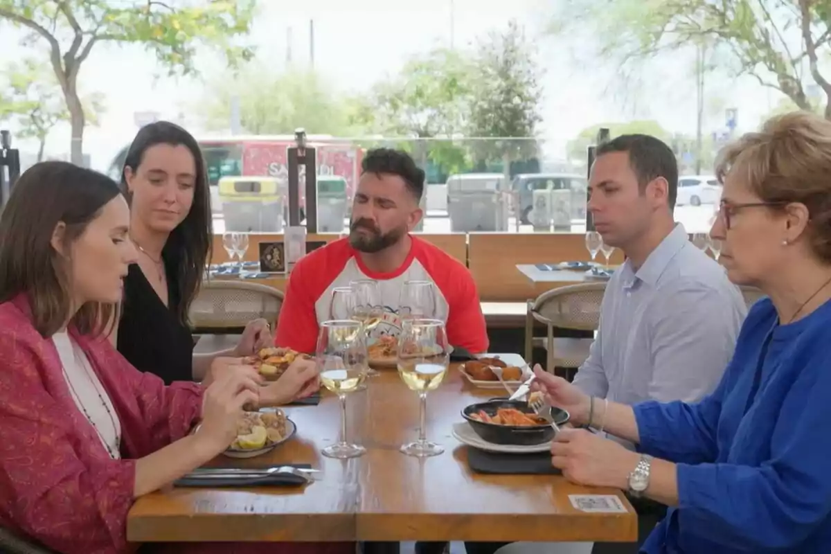 Un grup de persones estan asseguts al voltant d'una taula en un restaurant a l'aire lliure, gaudint d'un àpat i conversant.