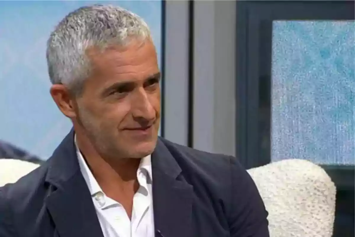 Hombre de cabello canoso con chaqueta oscura y camisa blanca sonriendo en un entorno interior.