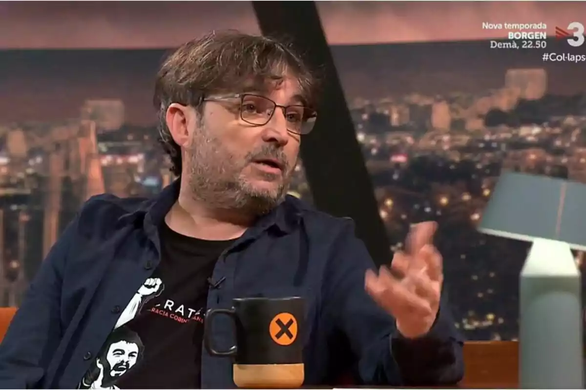 Un hombre con gafas y barba está sentado en un set de televisión sosteniendo una taza negra con un diseño naranja mientras habla gesticulando con la mano.