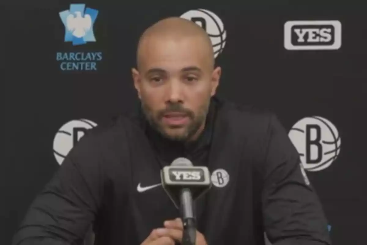 Un hombre calvo con barba está hablando en una conferencia de prensa, con un micrófono que tiene el logo de "YES" y un fondo con logotipos del Barclays Center y un equipo de baloncesto.