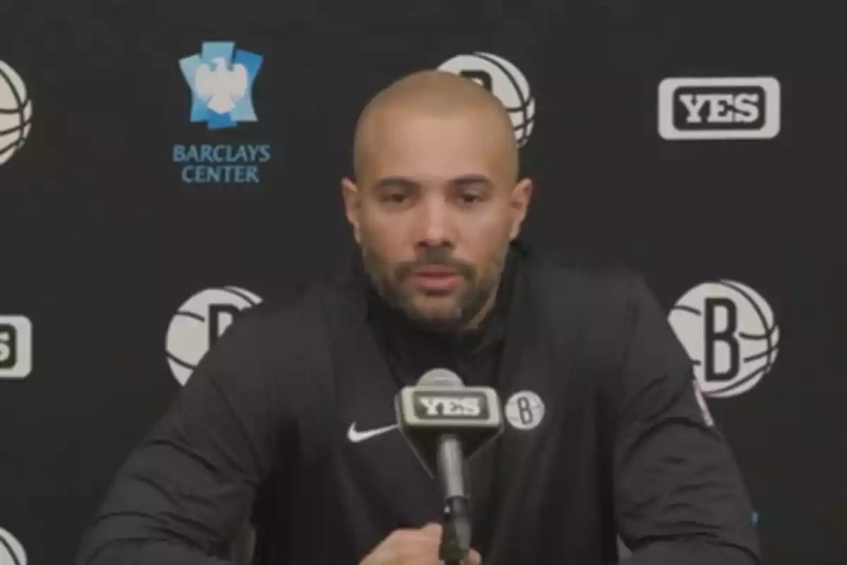 Un hombre calvo con barba en una conferencia de prensa con un micrófono frente a él y logotipos de Barclays Center y YES en el fondo.