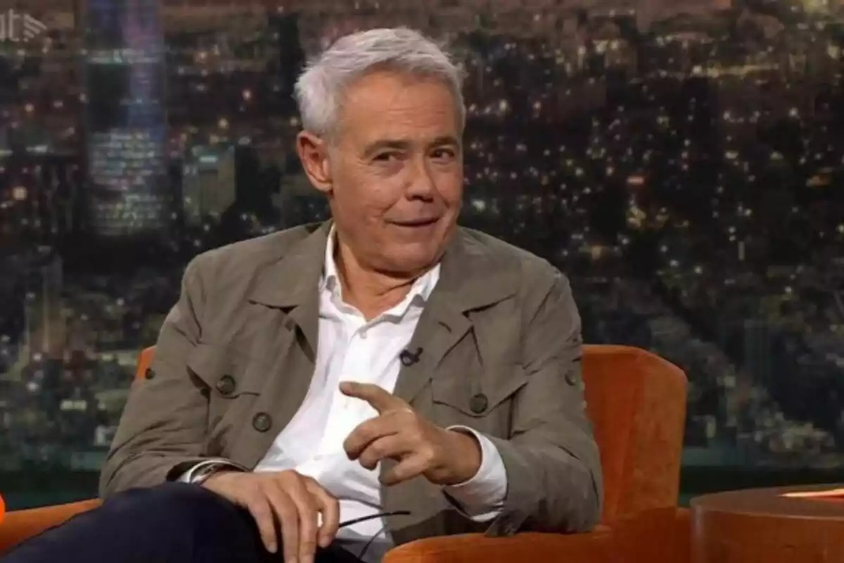 Home de cabells canosos assegut en un sofà taronja durant una entrevista en un plató de televisió amb un fons de ciutat il·luminada Home de cabells canosos assegut en un sofà taronja durant una entrevista en un plató de televisió amb un fons de ciutat il·luminada