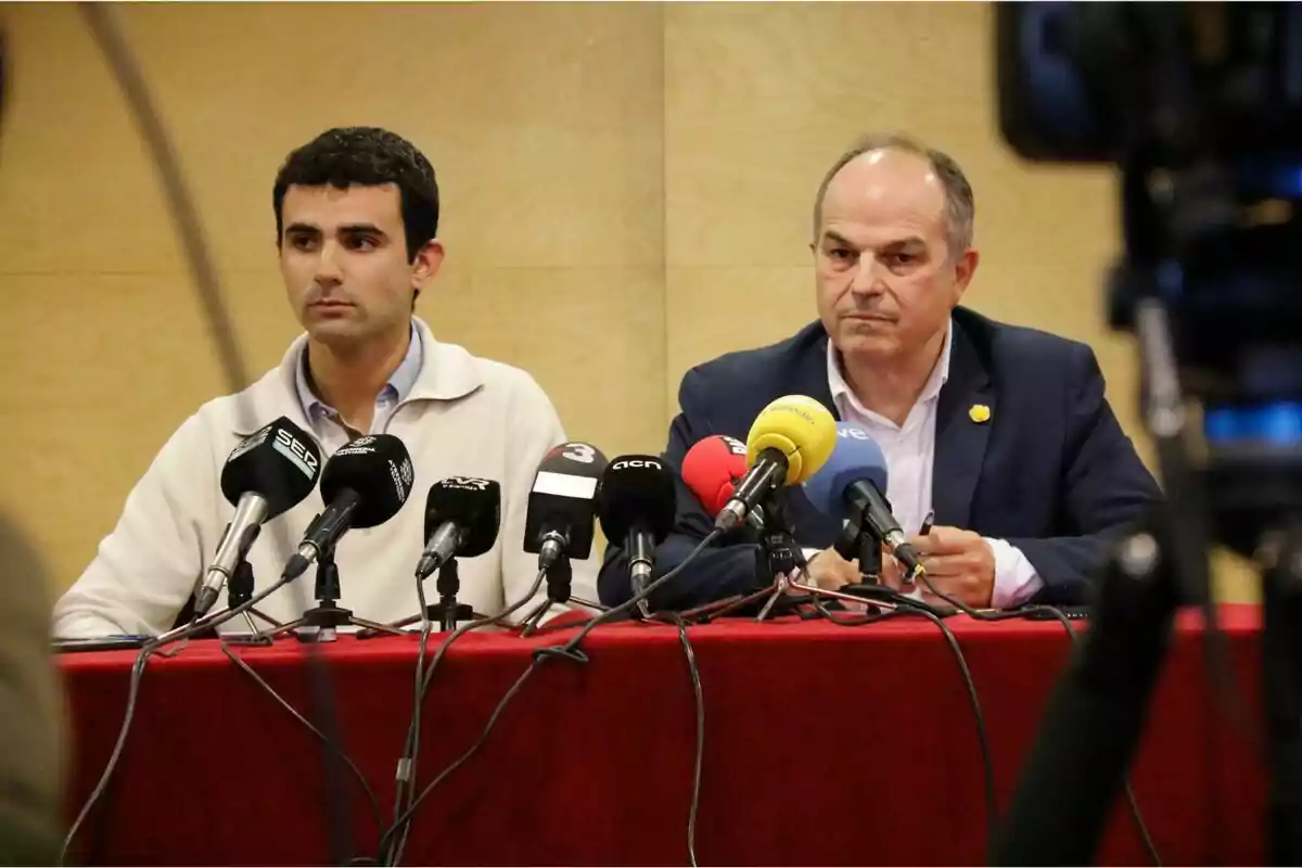 Dos hombres sentados detrás de una mesa con varios micrófonos durante una conferencia de prensa.