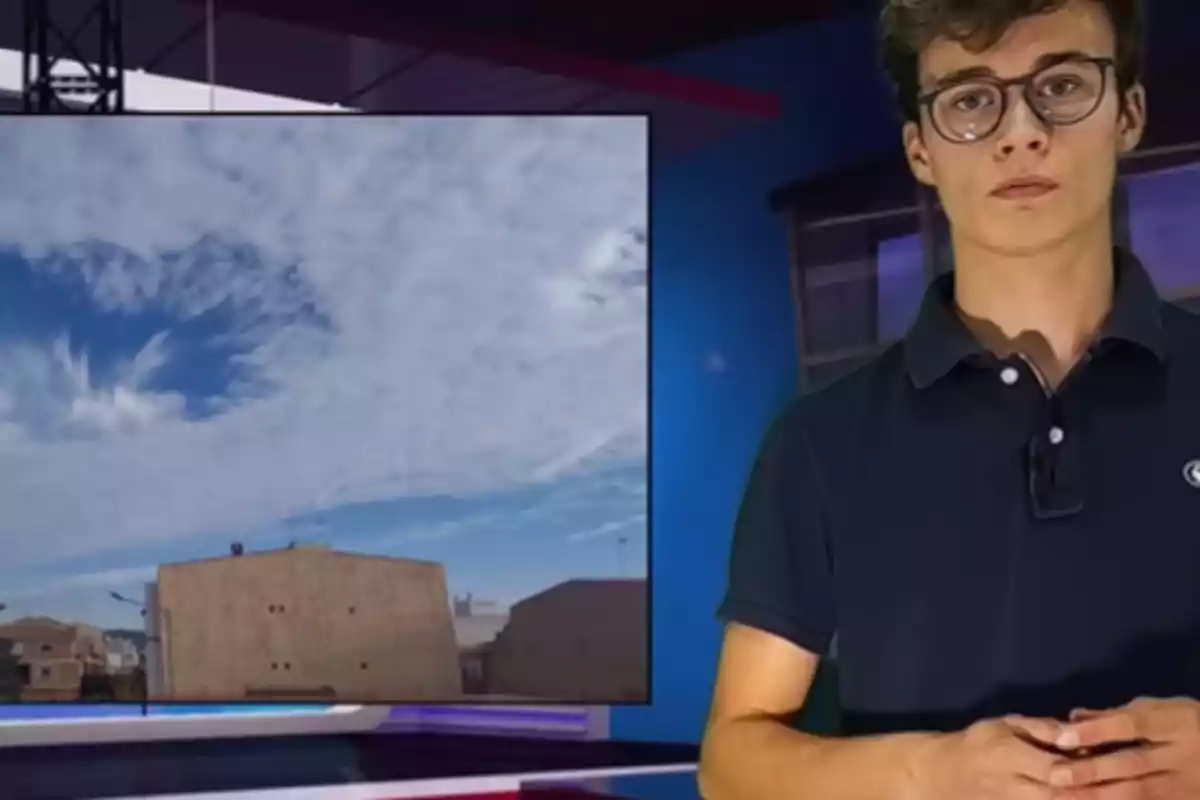 Un joven con gafas y camisa oscura presenta el clima en un estudio de televisión con una imagen de nubes en el cielo proyectada a su lado.