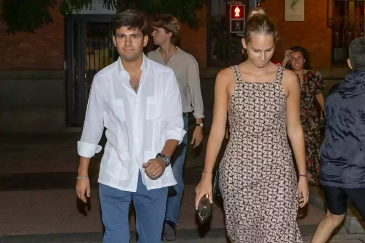 Un grupo de personas caminando por la calle durante la noche, con un hombre de camisa blanca y una mujer de vestido estampado al frente. Un grupo de personas caminando por la calle durante la noche, con un hombre de camisa blanca y una mujer de vestido estampado al frente.