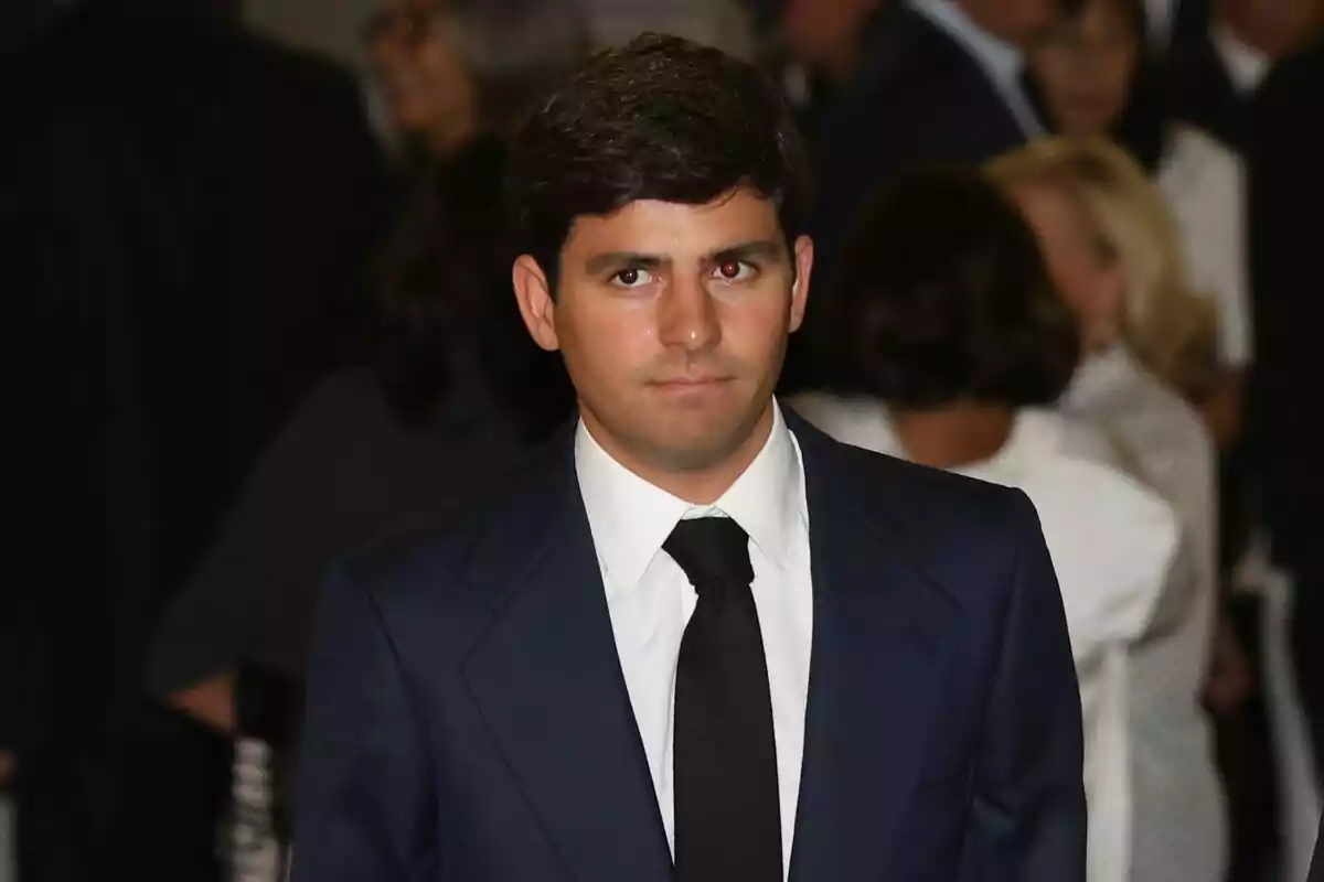 Pla curt de Juan Urquijo amb cabell fosc i curt, vestit amb un vestit blau marí, camisa blanca i corbata negra, està dret en un entorn amb diverses persones al fons. Pla curt de Juan Urquijo amb cabell fosc i curt, vestit amb un vestit blau marí, camisa blanca i corbata negra, està dret en un entorn amb diverses persones al fons.