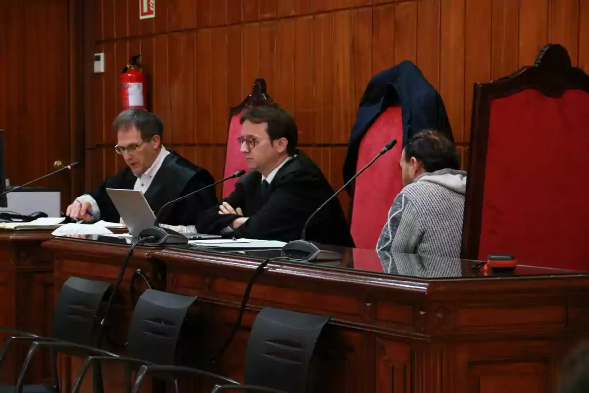 Tres persones assegudes en una taula de fusta en una sala de vistes, dues d'elles amb toga i una amb roba casual, amb micròfons i documents davant d'elles. Tres persones assegudes en una taula de fusta en una sala de vistes, dues d'elles amb toga i una amb roba casual, amb micròfons i documents davant d'elles.