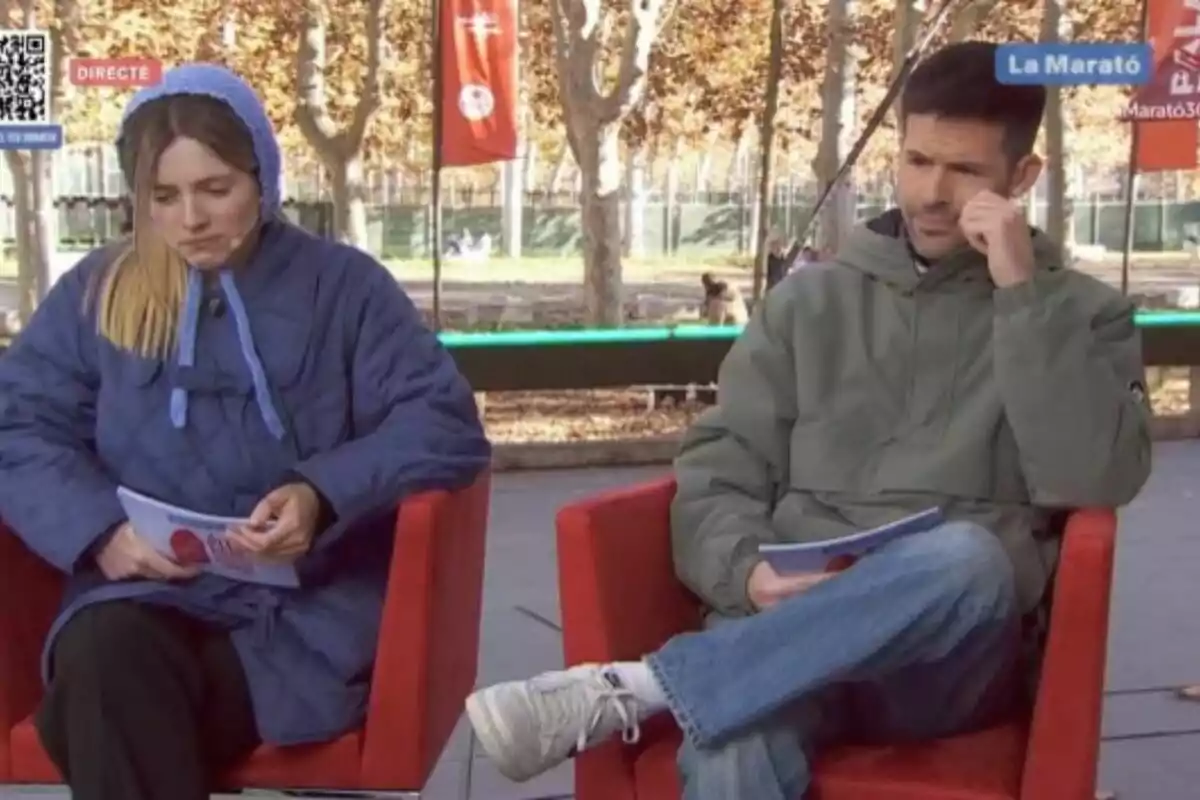 Dos personas sentadas en sillas rojas al aire libre durante una transmisión en vivo.