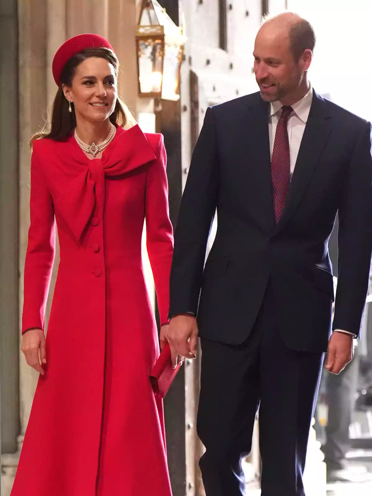 Kate Middleton y el príncipe Guillermo elegantemente vestida camina de la mano en un evento formal, ella lleva un vestido rojo con un sombrero a juego y él viste un traje oscuro con corbata. Kate Middleton y el príncipe Guillermo elegantemente vestida camina de la mano en un evento formal, ella lleva un vestido rojo con un sombrero a juego y él viste un traje oscuro con corbata.