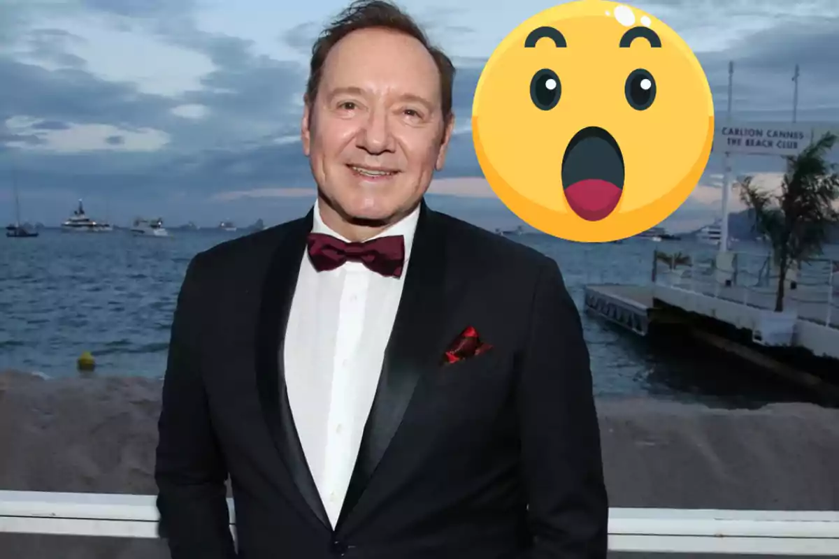 Un home amb esmòquing i llacet vermell somriu davant del mar amb un emoji sorprès superposat.