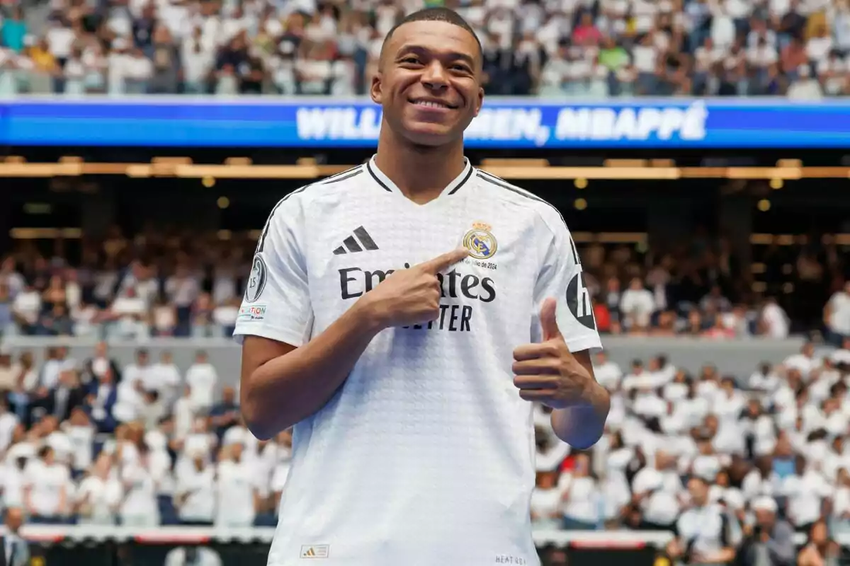 Jugador de fútbol con camiseta blanca del Real Madrid sonriendo y señalando el escudo del equipo en un estadio lleno de aficionados.