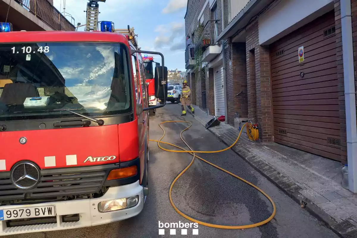 Un camió de bombers vermell està estacionat en un carrer estret al costat d'un edifici de maó, amb mànegues esteses a terra i un bomber caminant cap al fons.