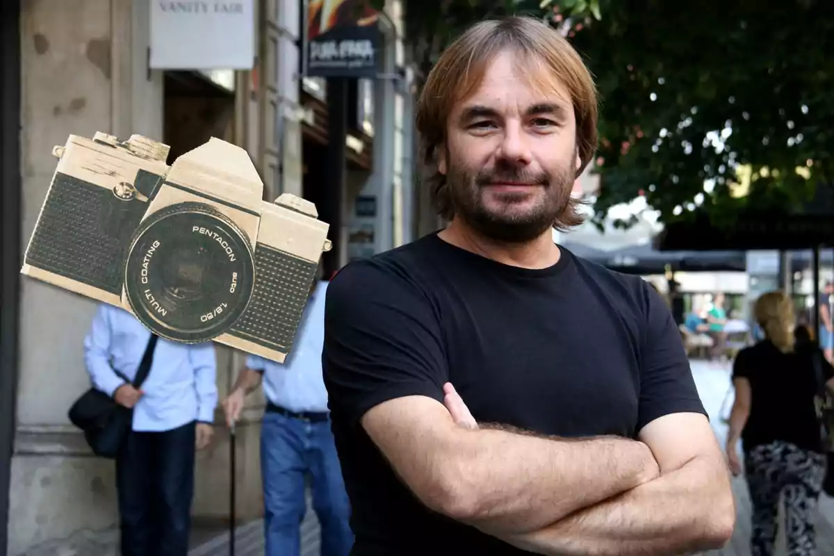 Un home amb barba i cabell llarg, vestit amb una samarreta negra, està dret amb els braços plegats en un carrer concorregut; a la cantonada superior esquerra de la imatge hi ha una càmera fotogràfica antiga superposada.