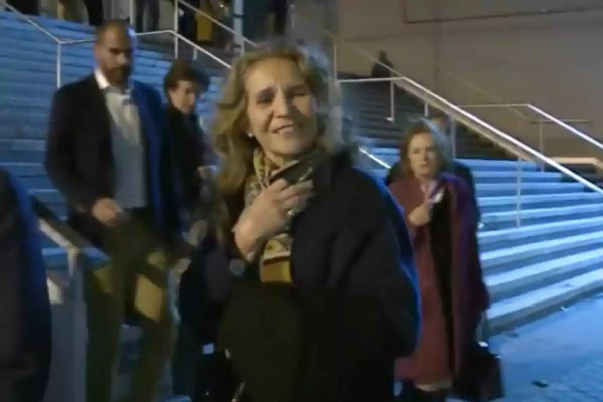 Una dona somrient amb una bufanda i un abric fosc està en primer pla, mentre que diverses persones caminen darrere seu en una escala il·luminada. Una dona somrient amb una bufanda i un abric fosc està en primer pla, mentre que diverses persones caminen darrere seu en una escala il·luminada.