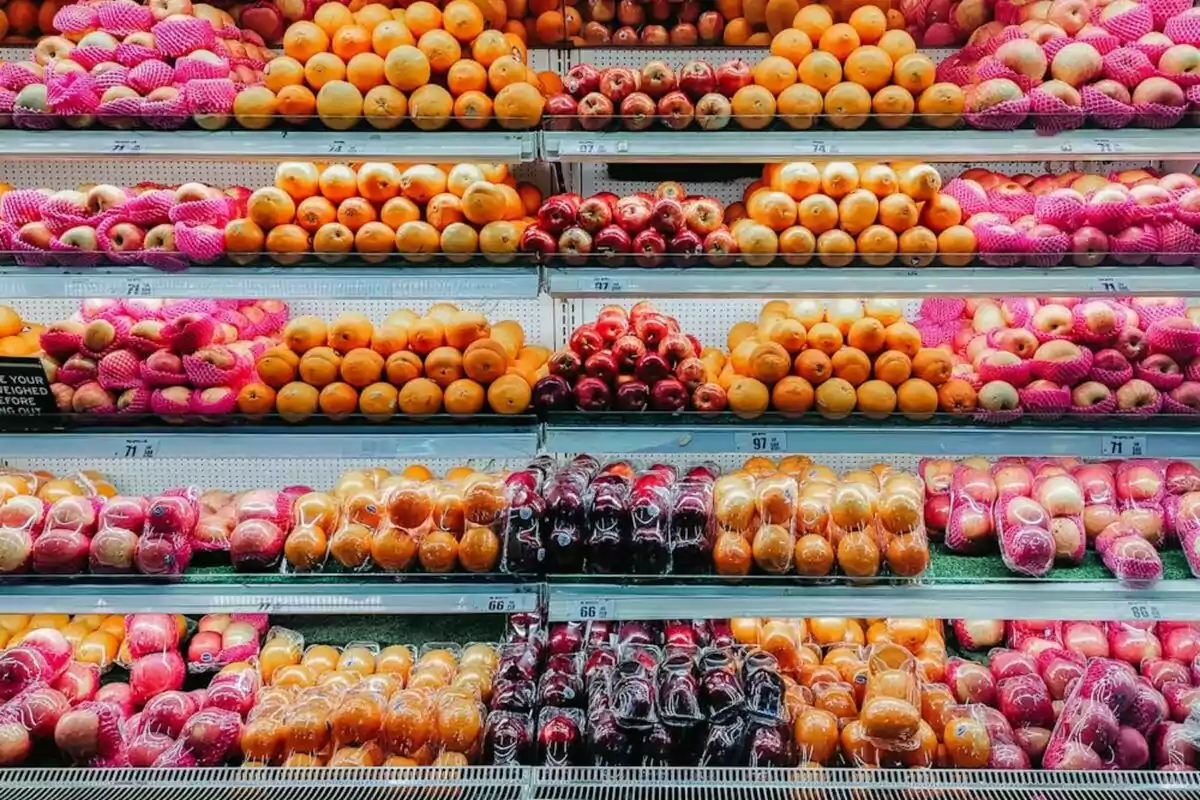 Estantes de supermercado llenos de frutas variadas como manzanas, naranjas y pomelos, algunas envueltas en redes protectoras rosas.