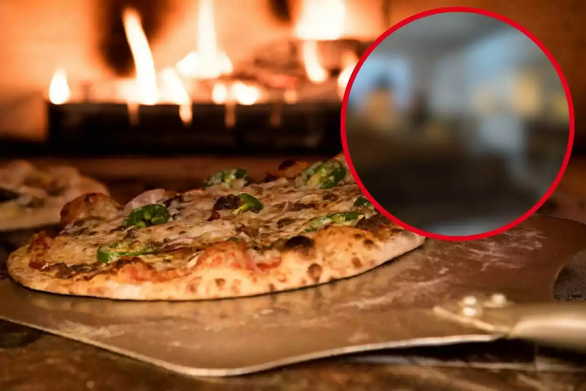 Pizza recién horneada en una pala frente a un horno de leña con llamas, con un círculo borroso en la esquina superior derecha.