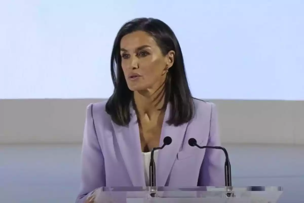 Mujer con cabello oscuro y lacio usando un saco lila hablando en un podio con dos micrófonos.