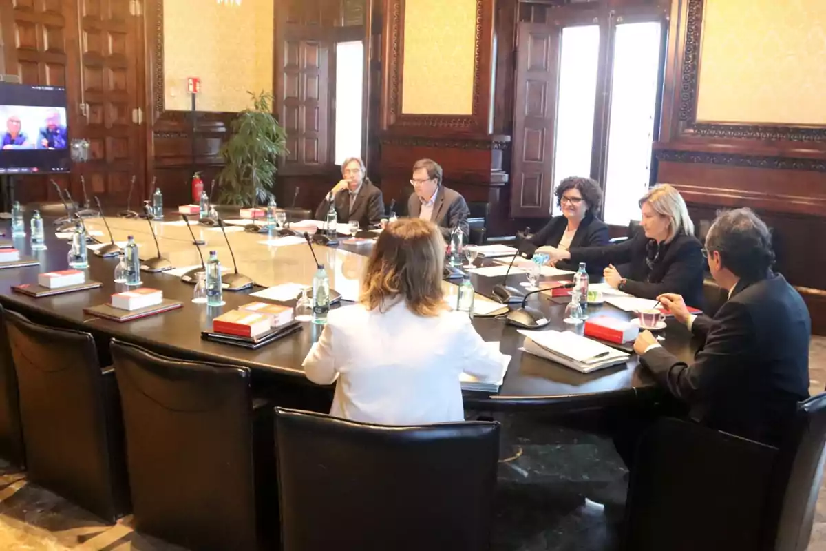 Un grupo de personas se encuentra reunido alrededor de una mesa ovalada en una sala de conferencias elegante, con libros y botellas de agua sobre la mesa y una pantalla mostrando una videollamada en el fondo.