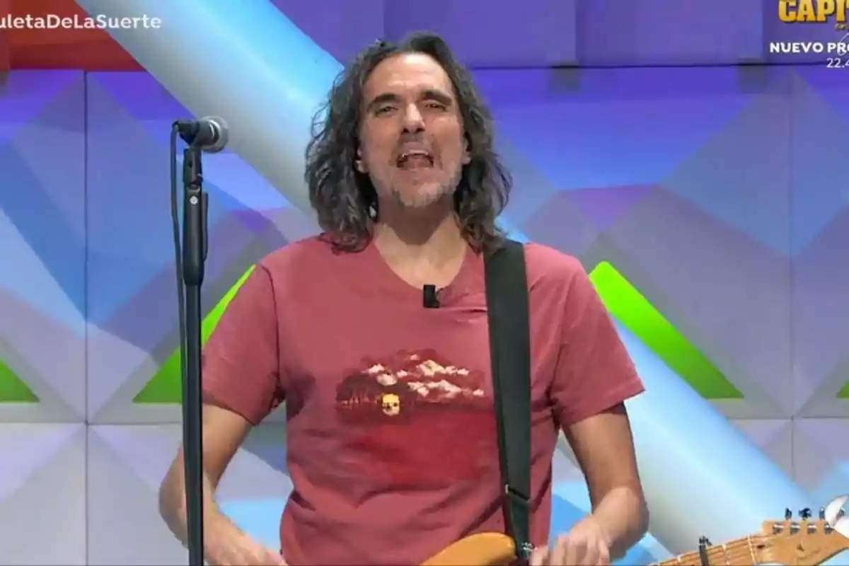 Joaquín Padilla con cabello largo y barba está de pie frente a un micrófono, vistiendo una camiseta roja con un diseño gráfico, en un escenario con fondo geométrico colorido. Captura de 'La Ruleta de la Suerte' Joaquín Padilla con cabello largo y barba está de pie frente a un micrófono, vistiendo una camiseta roja con un diseño gráfico, en un escenario con fondo geométrico colorido. Captura de 'La Ruleta de la Suerte'