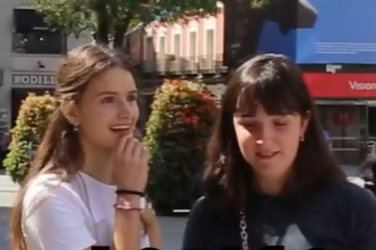 Dos personas jóvenes están de pie al aire libre en una calle con árboles y edificios al fondo, una de ellas sonríe mientras la otra tiene los ojos cerrados.