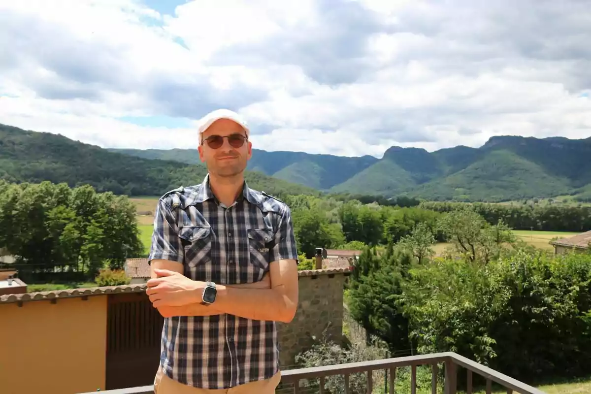 Un home amb ulleres de sol i gorra blanca, vestit amb una camisa de quadres, està dret amb els braços plegats en un balcó amb un paisatge muntanyós i verd de fons.