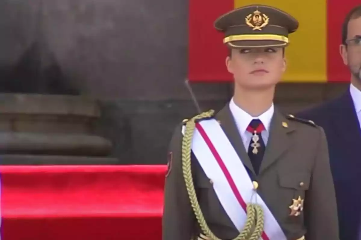 Una dona en uniforme militar amb una banda blanca i vermella, dreta davant una bandera.