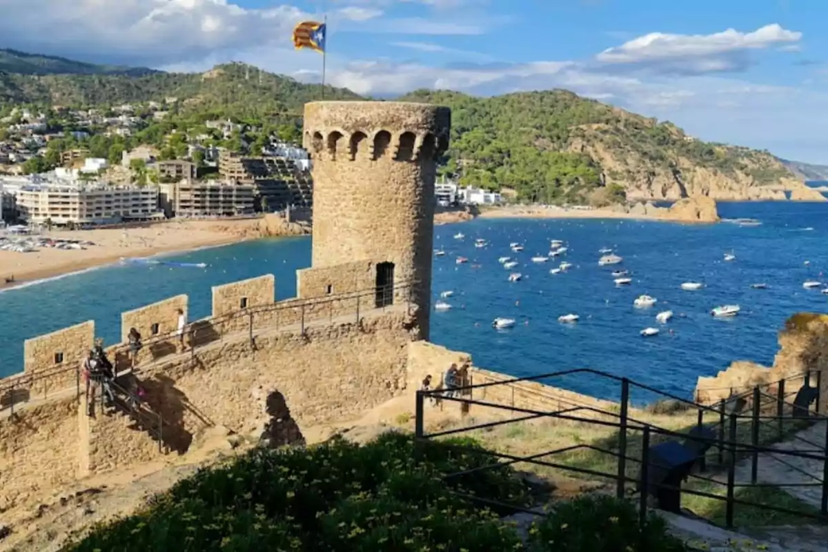 Vista d'una torre de pedra amb una bandera al cim, ubicada en una fortalesa costanera amb un mar blau i diverses embarcacions, i una ciutat amb edificis i turons verds al fons. Vista d'una torre de pedra amb una bandera al cim, ubicada en una fortalesa costanera amb un mar blau i diverses embarcacions, i una ciutat amb edificis i turons verds al fons.
