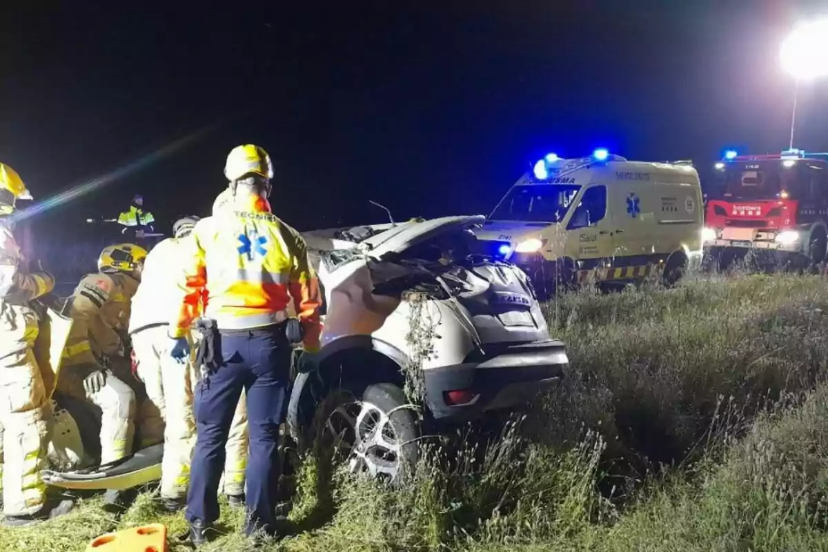 Un grupo de rescatistas y personal de emergencia atiende un accidente automovilístico nocturno en una carretera, con ambulancias y un camión de bomberos iluminando la escena.