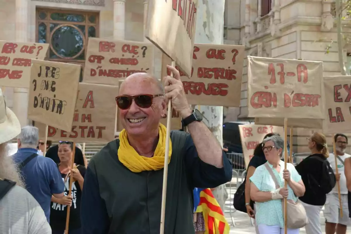 Un grup de persones participa en una manifestació sostenint pancartes amb missatges escrits, mentre que un home somrient amb ulleres de sol i una bufanda groga destaca en primer pla.