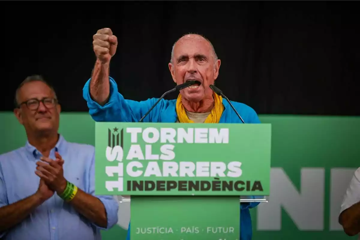 Home parlant en un míting amb un cartell verd que diu Tornem als carrers independència i una altra persona aplaudint al fons.