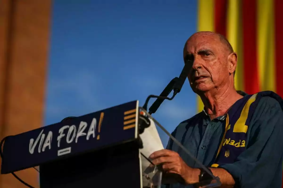 Un hombre hablando en un podio con un micrófono y una bandera de fondo.