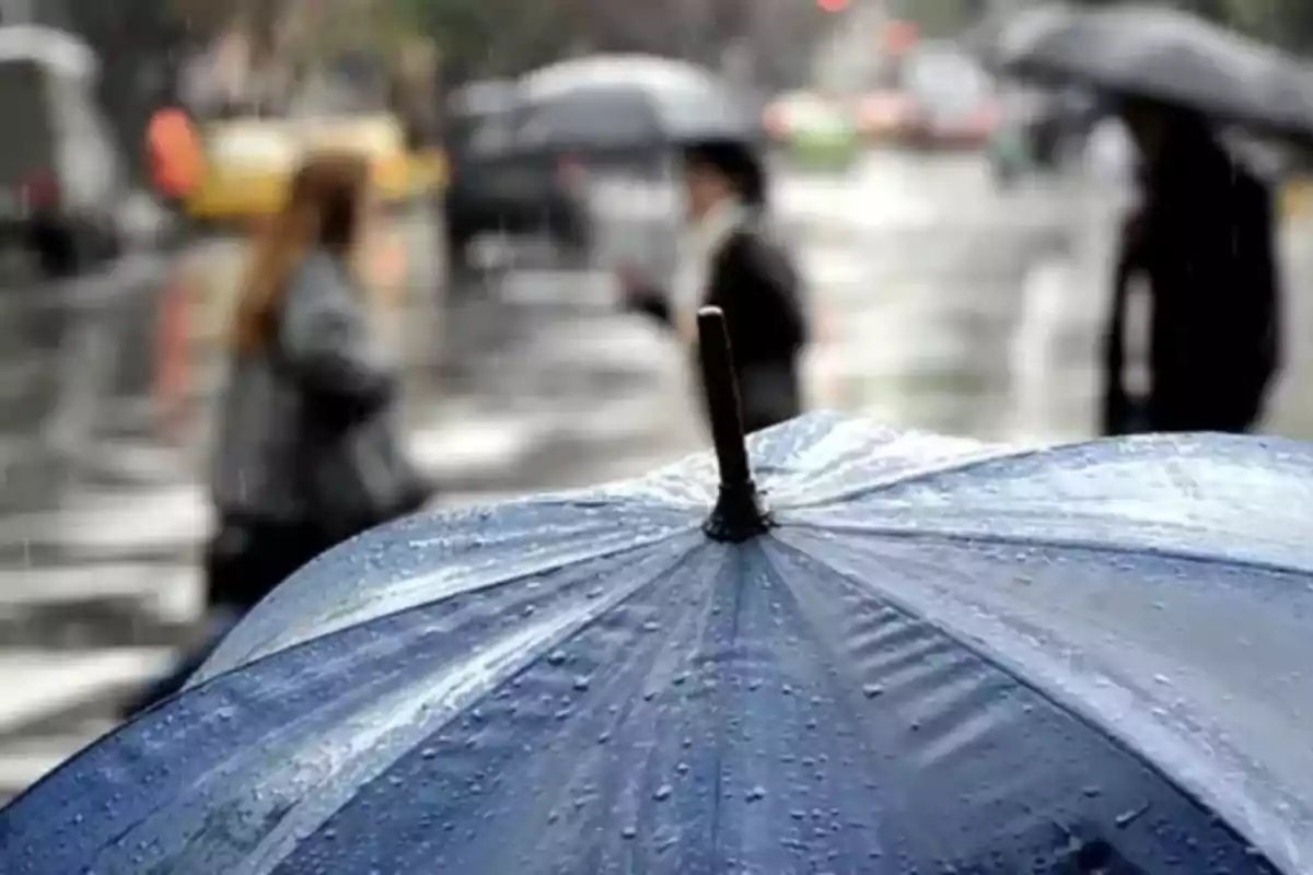 Un paraigua blau amb gotes de pluja en primer pla i persones desenfocades caminant sota la pluja al fons.