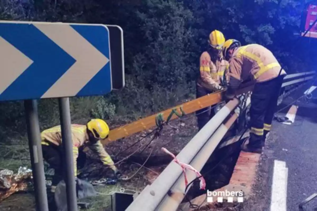 Bomberos trabajando en un rescate junto a una carretera con una señal de tráfico y barandilla metálica.