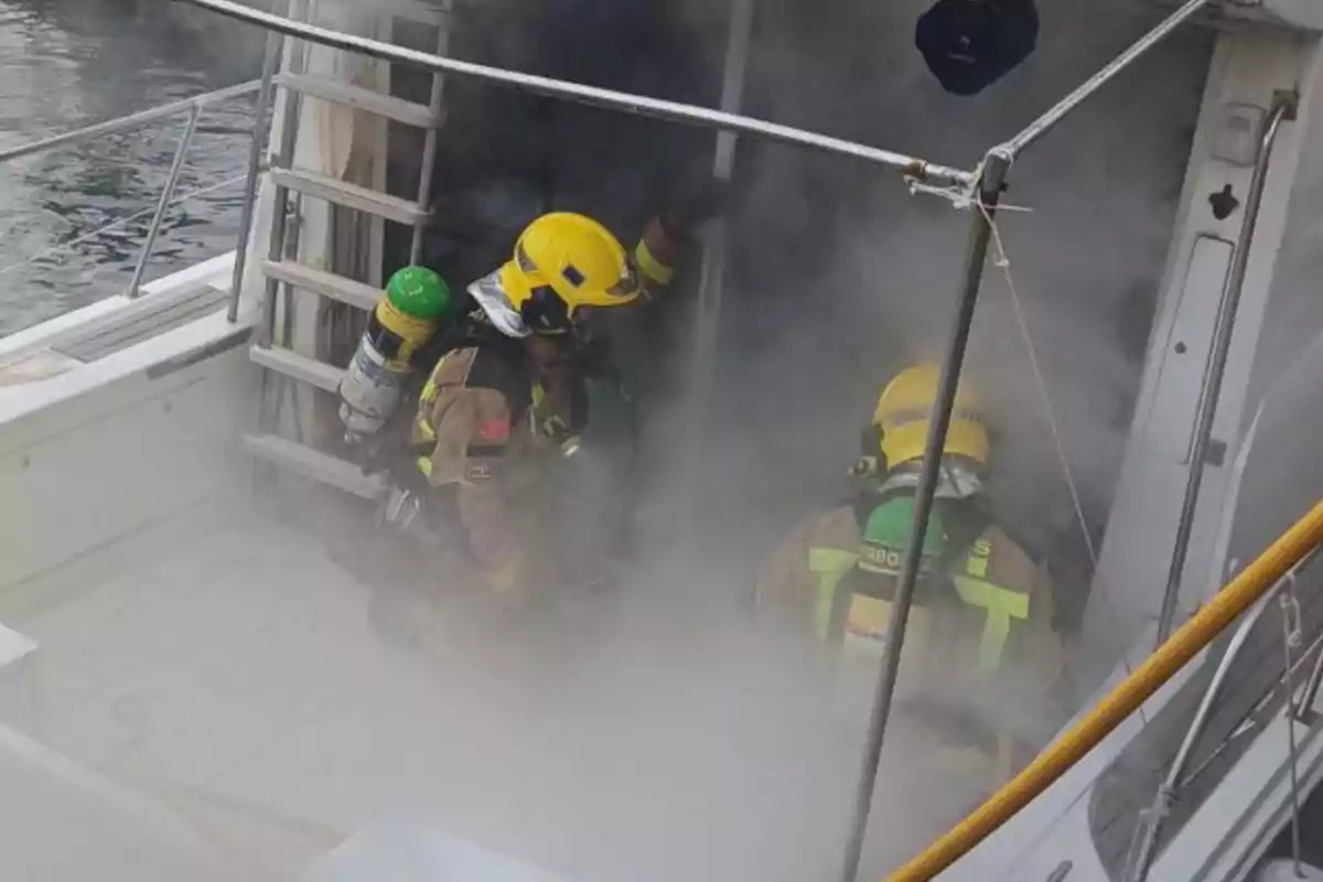 Bomberos con equipo de protección y cascos amarillos trabajando en un barco rodeado de humo.