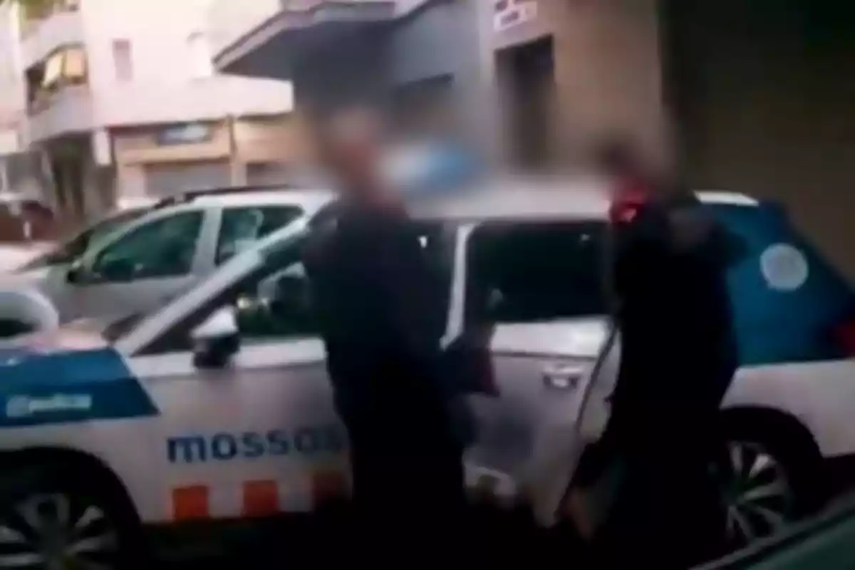 Dues persones dempeus al costat d'un cotxe de policia en un carrer urbà.