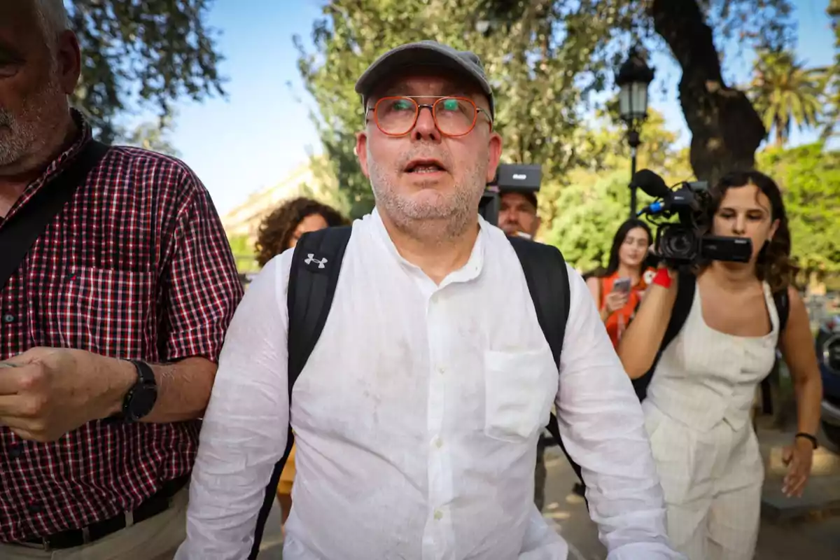 Un home amb ulleres i gorra, vestit amb una camisa blanca i motxilla, envoltat de persones i càmeres en un entorn exterior amb arbres.