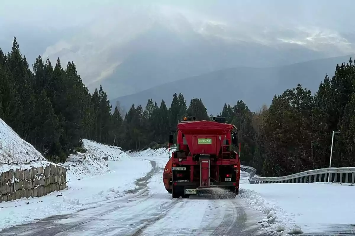 Un camió llevaneu vermell aclareix una carretera coberta de neu envoltada d'arbres i muntanyes.